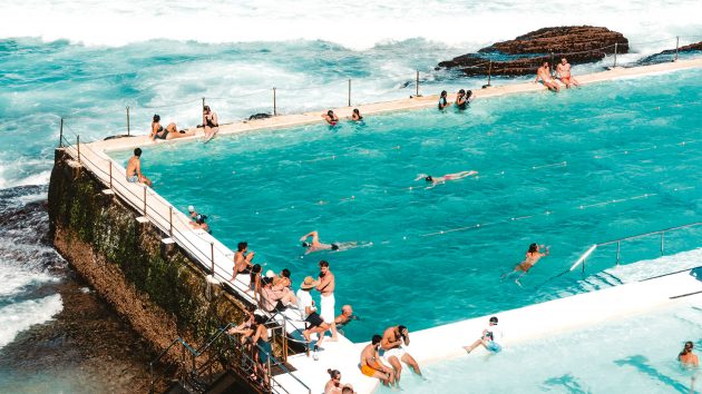 Rectangular outside swimming pool next to the waves. The sun is shining and many people are swimming and lounging.